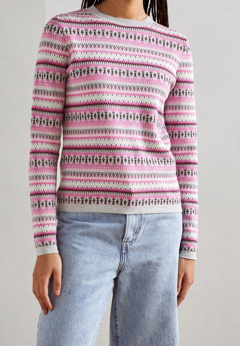 Pull long-sleeve gris clair avec un motif géométrique rose et noir, col et poignets côtelés, associé à un jean en denim bleu clair.