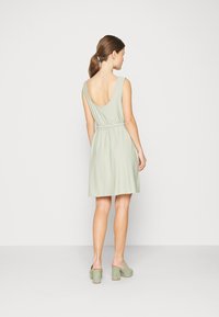 Vestido verde claro com decote nas costas, cintura cingida e saia expandida. Combinado com mules de salto verde claro, textura de tecido suave.