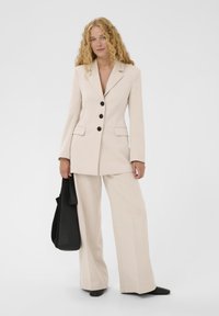 Blazer beige sur mesure avec boutons noirs, associé à un pantalon beige à jambes larges. Le mannequin tient un sac fourre-tout noir. Texture lisse, design structuré.