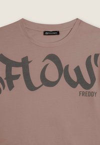 Camiseta de algodón rosa con un gran texto gráfico en gris oscuro "FLOW" con tipografía estilizada y una pequeña etiqueta de la marca "FREDDY" a un lado.