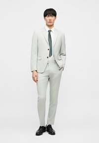 Νέος άνδρας που στέκεται φορώντας ανοιχτογκρι κοστούμι, λευκό πουκάμισο, πράσινη γραβάτα με σχέδια και μαύρα επίσημα παπούτσια σε απλό λευκό φόντο.