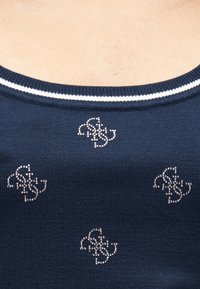 Μπλε ναυτικό πλεκτό τοπ με ασημένια σχέδια από στρας και λευκή ριγέ λαιμόκοψη. Η υφή είναι απλή και η εφαρμογή στενή.