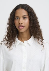 Hvid bluse med en skjortekrave med knapper, prydet med et subtelt polkaprikket mønster og en let, glat tekstur.