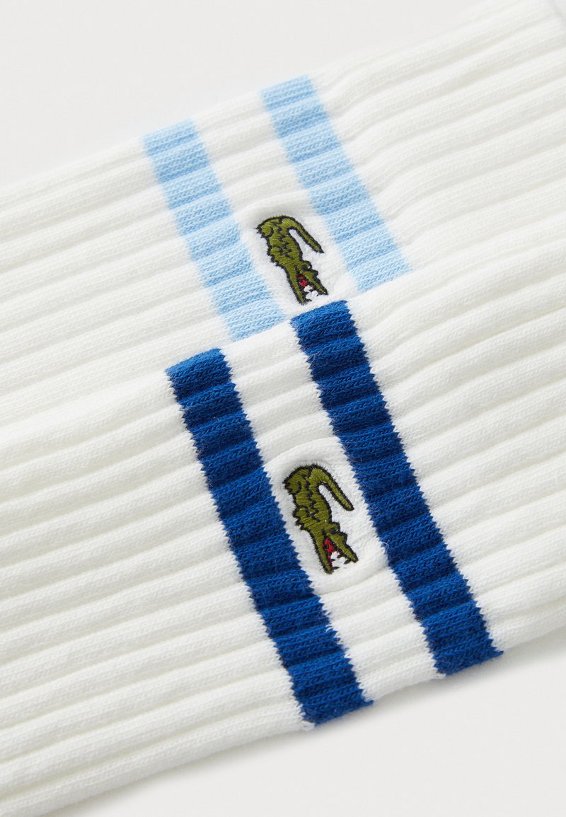 Chaussettes blanches côtelées avec des rayures bleues et bleu clair. Logo de crocodile vert brodé entre les rayures. Texture de tissu douce.