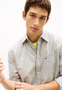 Νέος άνδρας με κοντά καστανά μαλλιά, φορώντας ένα ανοιχτό πράσινο και λευκό πουκάμισο με καρό κουμπιά πάνω από ένα νέον πράσινο μπλουζάκι, σε λευκό απλό φόντο.