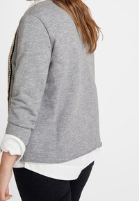 Grauer Sweatshirt mit strukturierter Oberfläche, verfügt über lange Ärmel, einen abgerundeten Saum und ist mit einem weißen Hemd darunter geschichtet.