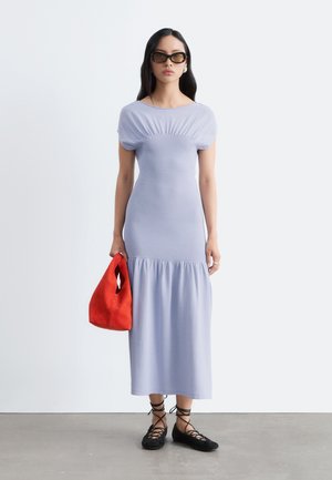 Femme portant une robe midi violette claire et des ballerines noires à lacets, tenant un sac rouge et portant des lunettes de soleil foncées sur un fond uni.