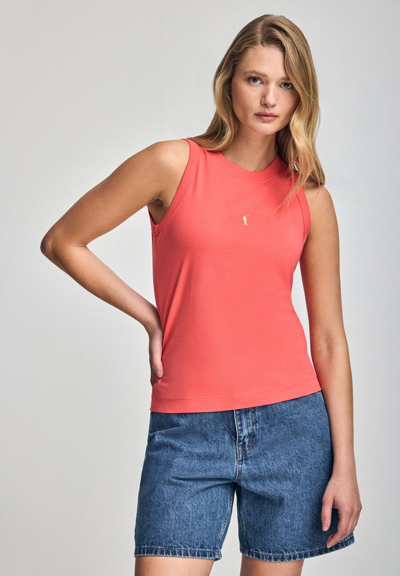 Koronkowy, bezrękawnik w kolorze koralowym, wykonany z gładkiego materiału, z okrągłym dekoltem i haftowanym logo. W zestawieniu z dżinsowymi szortami, które prezentują luźny krój.