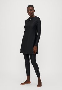 Juodas sportinis tunikas ilgomis rankovėmis, pagamintas iš lygaus audinio, derinamas su juodais leggingais, kurių kulkšnį puošia baltas Nike logotipas.