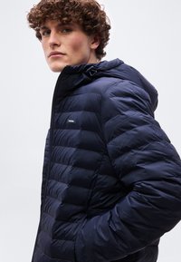 Νεαρός με σγουρά μαλλιά που φοράει μπλε ναυτικό καπιτονέ μπουφάν με κουκούλα, τα χέρια στις τσέπες, κοιτάζοντας προς τα αριστερά.