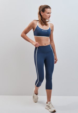Giovane donna con top sportivo blu navy e leggings con strisce bianche, in posa a metà passo su uno sfondo grigio chiaro uniforme.