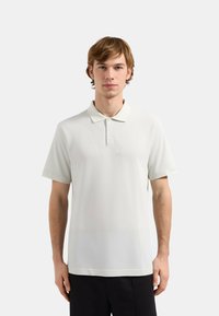 Polo shirt beige chiaro con colletto, maniche corte e dettagli di cucitura sottili. Presenta un logo sulla manica sinistra. Texture liscia.
