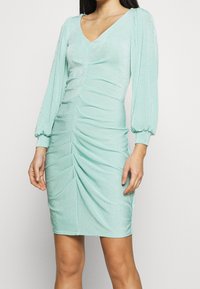 Robe vert menthe avec un col en V, des manches bouffantes longues et des détails froncés à l'avant. Fabriquée dans un tissu doux et brillant avec une silhouette ajustée.