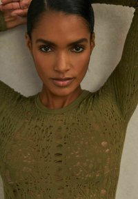 Olívazöld, texturált anyagból készült felső, bonyolult kivágott mintával, hosszú ujjakkal és kerek nyakkivágással.