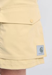 Geltonas iki kelių pasiekiantis sijonas, pagamintas iš lygaus audinio, su priekinėmis kišenėmis, užsifiksuojančiomis spaustuku, ir etikete su logotipu.