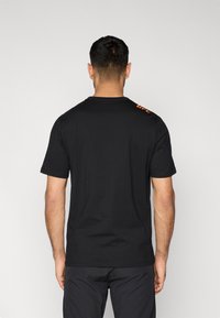 Μαύρο βαμβακερό μπλουζάκι με κοντά μανίκια, στρογγυλή λαιμόκοψη και πορτοκαλί λογότυπο UFC στον ώμο. Λεία υφή, κανονική εφαρμογή.