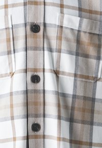 Rutet skjorte i hvit, beige og grå. Har en myk tekstur, to brystlommer og svarte knapper foran.