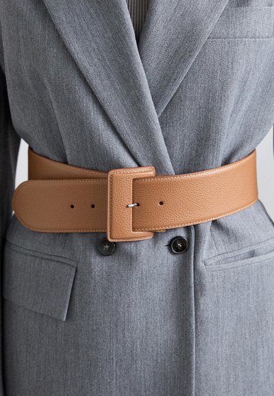Ceinture en cuir tan avec une texture lisse et boucle rectangulaire, portée sur un blazer grise sur mesure avec un motif vertical subtil.