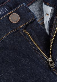 Tumši zili denim džinsi ar redzamu rāvējslēdzēju un vara pogu. Iekļauj zelta diegu un auduma etiķeti jostasvietā.