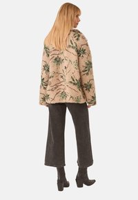 Chaqueta beige difusa con bordados florales verdes y lentejuelas, llevada sobre pantalones de pierna ancha oscuros y botas de tacón negro. Vista trasera, tejido texturizado.