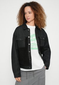 Fekete farmerruha, elöl bolyhos fekete panellel, patentos gombokkal, két mellzsebbel és laza szabással. Egy grafikás póló fölött viselve.