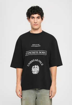 Young man with curly hair wears oversized black T-shirt with white text: "Kids of the loud kingdom," "Concrete born," and "Unbreakable" emblem.