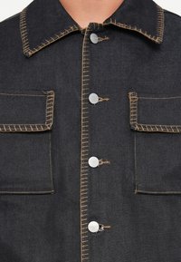 Donkerdenim jacket met zilveren knopen, contrasterende tan stiksel, twee borstzakken en een klassiek kraagontwerp.