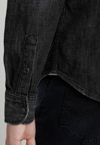 Svart denimjacka med knäppt ärmslut, med en något sliten fåll. Tyget har en strukturerad yta med ett subtilt mönster.