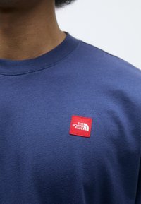 Tamsiai mėlynas medvilninis džemperis su apykakle, turintis raudoną kvadratinį logotipo patch'ą su tekstu "The North Face" ir baltu išsiuvinėjimu viršuje.