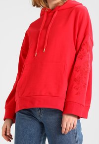 Sudadera con capucha roja con un bolsillo canguro, bordado floral texturizado en la manga izquierda y cordón ajustable. Tela suave, corte relajado.