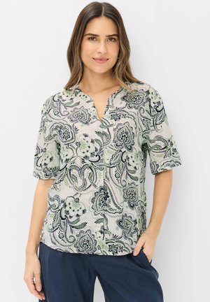 Frau trägt eine kurzärmelige Bluse mit V-Ausschnitt und schwarzen sowie grünen Paisley-Blumenmustern, kombiniert mit dunkelblauer Hose, steht mit einer Hand in der Tasche.