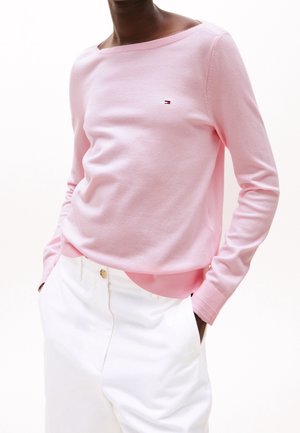 Personne portant un pull léger à manches longues rose clair avec un petit logo et un pantalon blanc taille haute, les mains dans les poches.