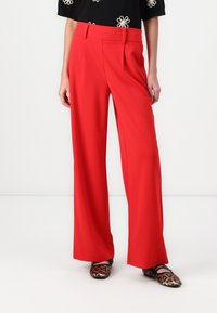 Pantalons larges rouges avec une texture lisse, des plis plats à l'avant et une taille cintrée, associés à des chaussures imprimées.