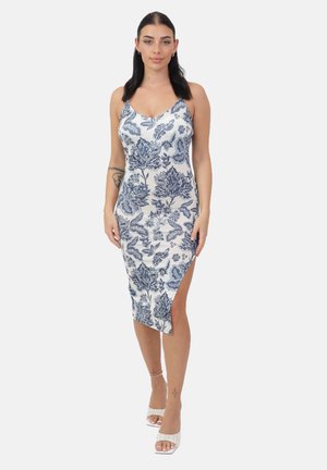 Vrouw draagt witte midi-jurk met blauw bloemenpatroon en een zijsplit, gecombineerd met witte sandalen met hak, staand tegen een effen achtergrond.