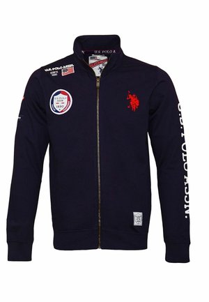 Marineblauwe jas met ritssluiting, met rood polo-spelerlogo, U.S. Polo Assn. badges en witte tekst op de rechter mouw.