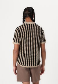 Maglione a strisce nero e beige a maniche corte, con design testurizzato, scollo rotondo e orlo a coste. Indossato con pantaloni corti marroni.