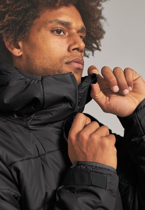 Man with curly hair zipping up a black puffer jacket collar, focusing on his face and hands against a plain background.