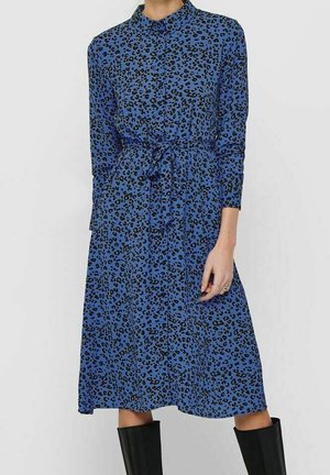 Robe midi bleue à manches longues avec imprimé léopard noir, col, fermeture boutonnée devant, ceinture à la taille, portée avec des bottes noires montantes jusqu’aux genoux.