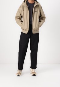 Veste beige à fermeture éclair avec capuche, associée à une chemise noire et un pantalon, ainsi qu'à des baskets beiges. Tissu texturé avec poignets et ourlet élastiques.