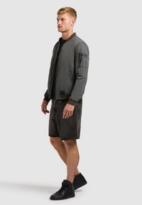 Homme portant une veste bomber grise, un T-shirt noir, un short en jean noir et des baskets montantes noires, debout de profil sur un fond uni.