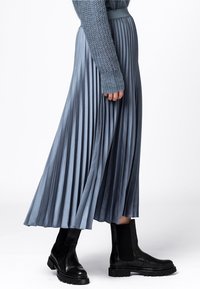 Plisserad ljusblå satinkjol med slät textur, tillsammans med svarta ankelboots, visad från sidan. Luksuriös drape och rörelse.