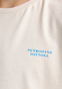 Camiseta de color crema claro hecha de suave algodón, con cuello redondo, y muestra el texto azul "PETROLIND DAYTONA".