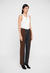 Weißes Rippen-Top mit gedrucktem Logo, kombiniert mit hochtaillierten, zweifarbigen braunen und dunkelgrauen Denim-Hosen. Schwarze Absatzpantoletten.