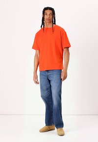T-shirt en coton orange avec une coupe décontractée et des manches courtes, associé à un jean droit bleu et des chaussures beiges. Design uni, sans motifs.