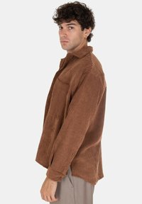 Camicia di velluto a coste marrone con una vestibilità comoda, caratterizzata da un colletto, maniche lunghe e una tasca frontale, mostrata di lato.