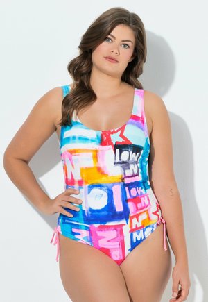 Žena s dlouhými hnědými vlasy, oblečená v barevných jednodílných plavkách s růžovými, modrými, oranžovými a grafickými textovými vzory, stojící na jednobarevném pozadí.