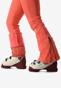 Skischoenen met een wit en bordeaux ontwerp, uitgerust met drie zwarte gespen en gemodelleerde zolen, gecombineerd met levendig koraalkleurige skibroek.