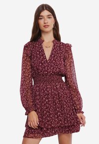 Robe florale bordeaux avec de longues manches transparentes, un col volanté et une taille smockée. Elle présente une jupe à volants et un motif floral subtil.