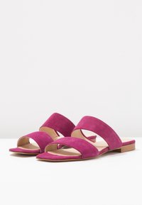 Pár fuchsiových semišových skládacích sandálů se dvěma širokými popruhy a nízkými dřevěnými podpatky na bílé ploše.