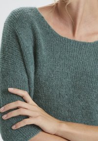 Zielony sweter dzianinowy z szerokim dekoltem i krótkimi rękawami. Miękka tekstura z prążkowanym wzorem. Dłoń oparta na rękawie.